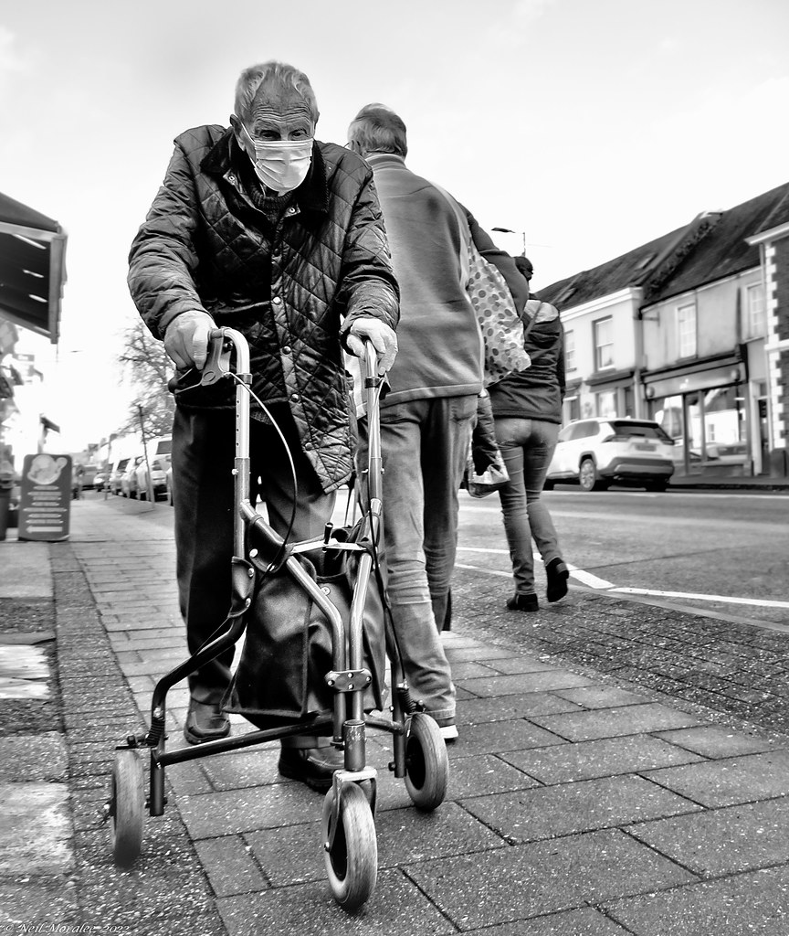 Black-and-white photo of an elderly masked man using a three-wheeled walker on a sidewalk. — illustrating “OSMO Girl Next Door Giftset” on Florence and Mary