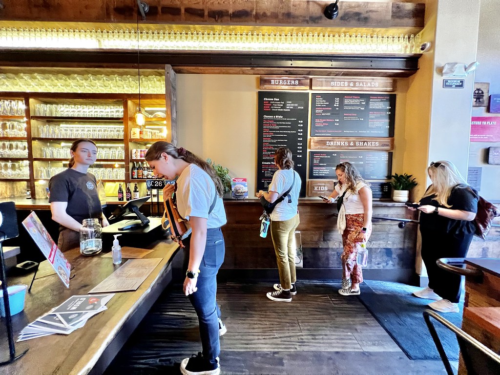 Customers line up at a rustic wooden restaurant counter with large wall menus and warm lighting. — illustrating “Patty & Bun Pilgrimage” on Florence and Mary