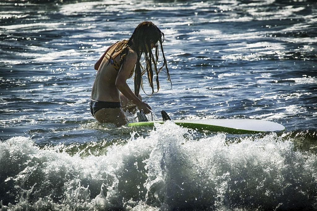 Woman with long dark hair holds an overturned white-green surfboard in blue ocean waves. — illustrating “Practice Makes Perfect” on Florence and Mary