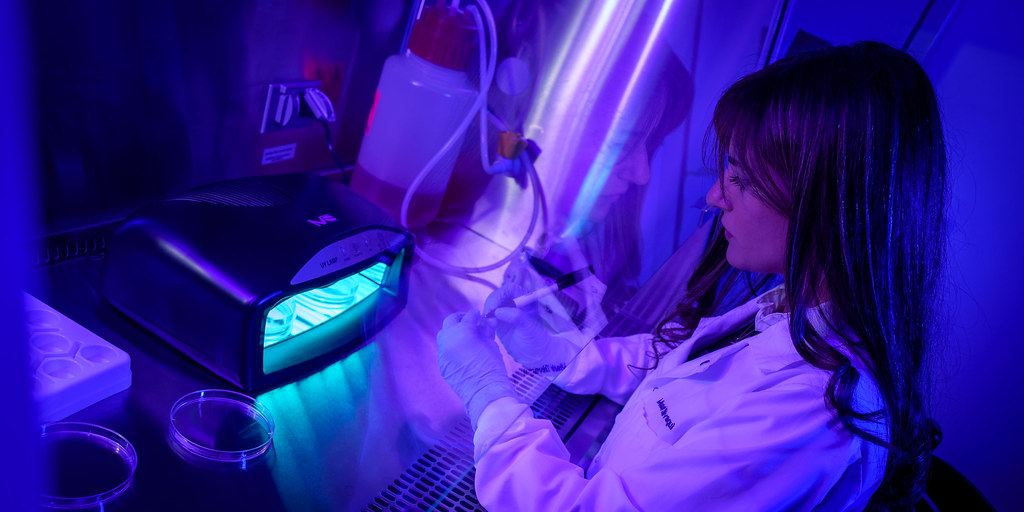 Woman in white lab coat and gloves works in a lab glowing with vibrant purple ultraviolet light. — illustrating “Product Fail | imPRESS Nails” on Florence and Mary