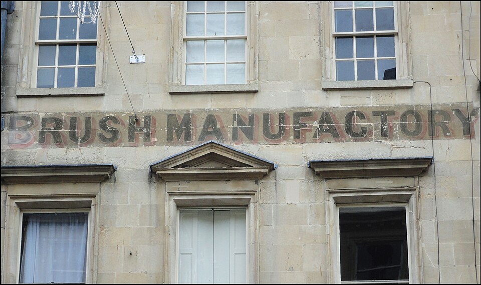 Weathered BRUSH MANUFACTORY lettering painted on a light stone building facade between windows. — illustrating “Review // Elle McPherson Rotating Dry Body Brush” on Florence and Mary