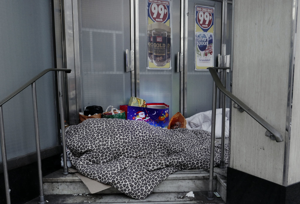 Person sleeps under leopard-print blanket in a shop doorway beside bags including a festive Santa bag. — illustrating “Sleep at Christmas” on Florence and Mary