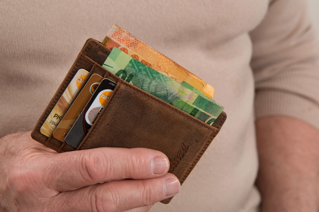 Hand holds weathered brown leather wallet with credit cards and colorful banknotes against neutral background. — illustrating “Spending Ban - Magazines” on Florence and Mary