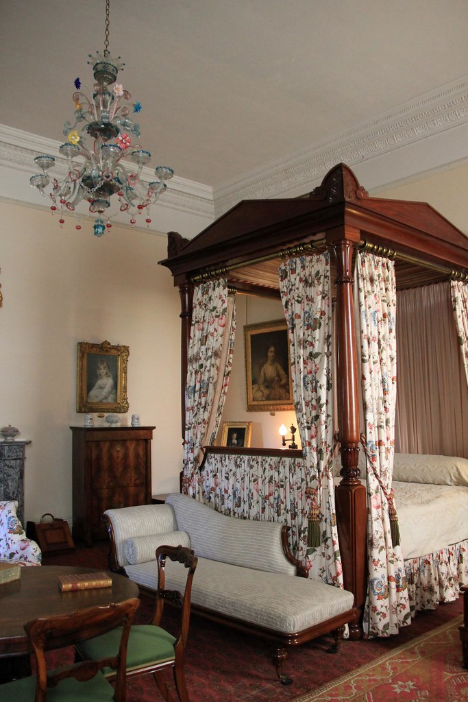 Ornate dark wood four-poster bed with floral curtains beside striped chaise under colorful chandelier. — illustrating “The Bedroom Redesign | In with the new” on Florence and Mary