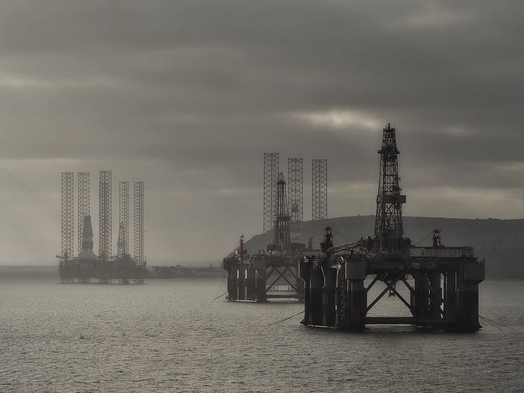 Moody desaturated photo of industrial offshore oil rigs with tall lattice towers in a dark calm sea. — illustrating “The British Tag” on Florence and Mary