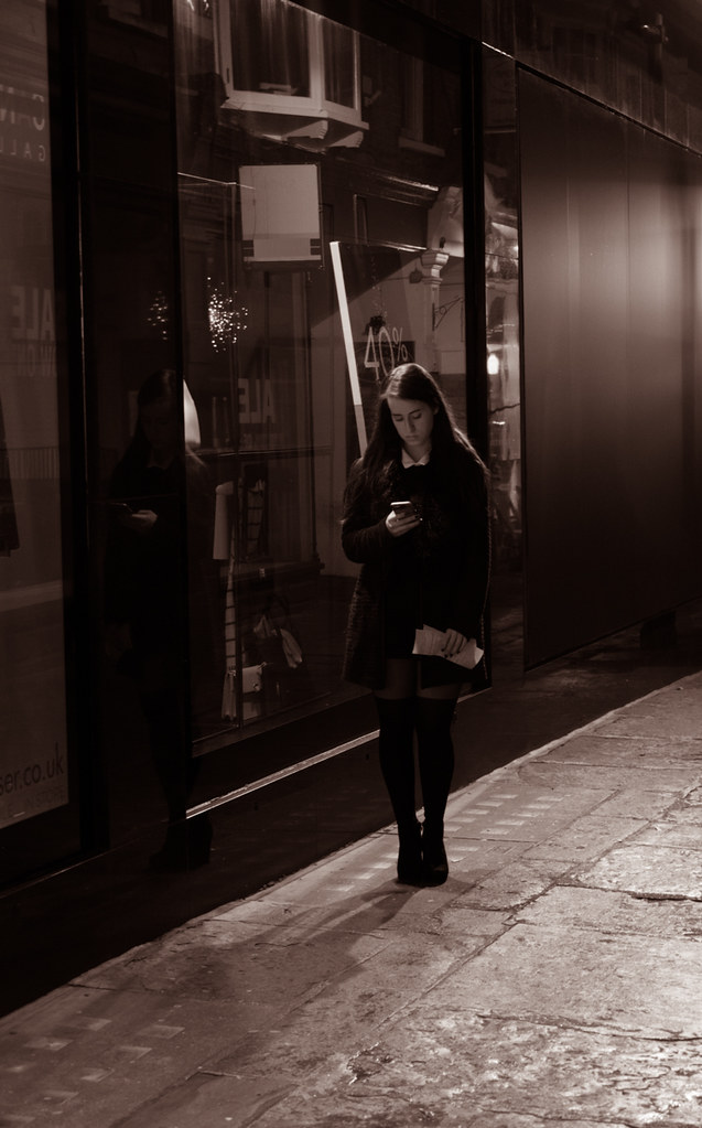 Sepia photo of a young woman on a night sidewalk looking at her phone before a storefront. — illustrating “The Single Girls Date Night #2” on Florence and Mary