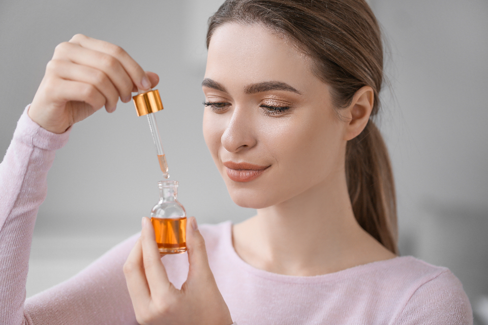 Woman with shimmering eyeshadow holds orange oil bottle and uses a dropper with a gold cap. — illustrating “The Skincare Tag” on Florence and Mary