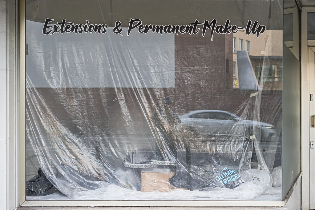 Shop window with Extensions and Permanent Make-Up signage draped in plastic reflecting a silver car. — illustrating “The Spending Ban” on Florence and Mary