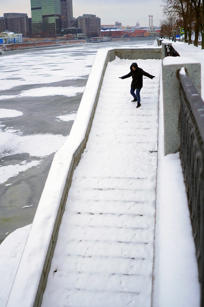 Person in a black hooded coat balances down snow-covered outdoor stairs beside a frozen urban river. — illustrating “The Winter Project // Winter Accessories” on Florence and Mary
