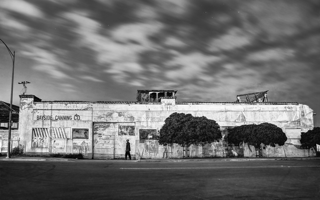 Black-and-white long exposure of someone walking past mural-covered Bayside Canning Co. under streaky sky. — illustrating “Two nights in San Francisco” on Florence and Mary