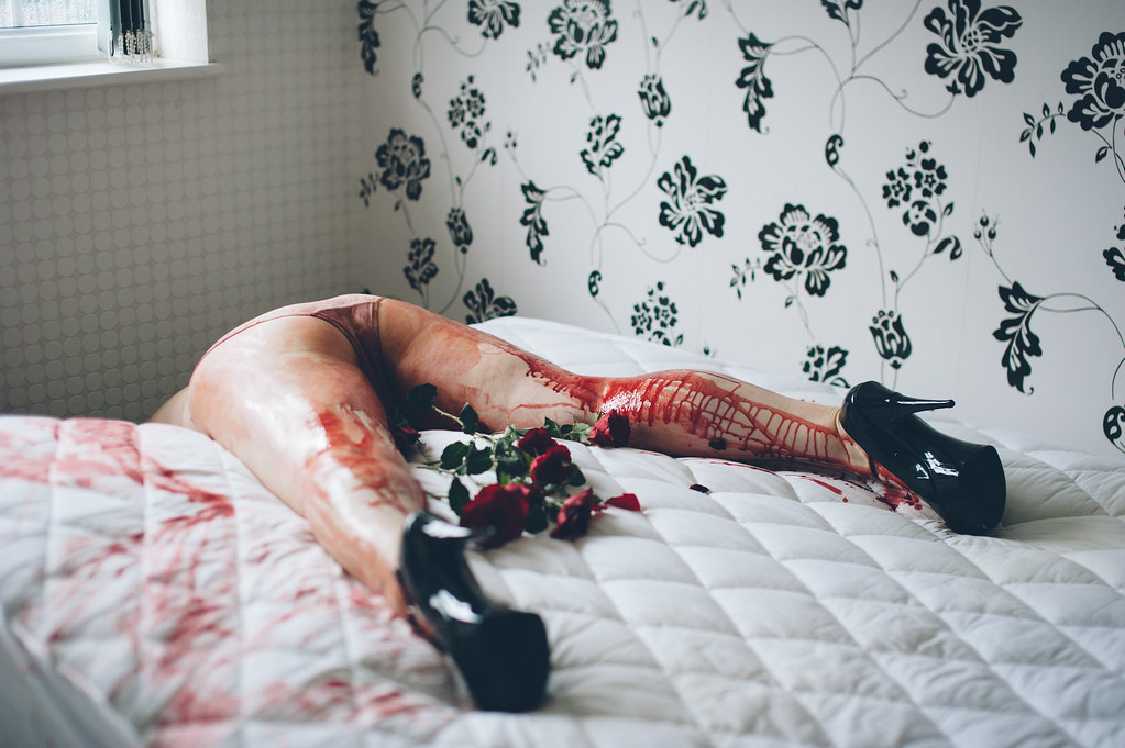 Red-stained legs in black high heels rest on a white quilted bed with roses and floral wallpaper. — illustrating “"V" Ladies” on Florence and Mary