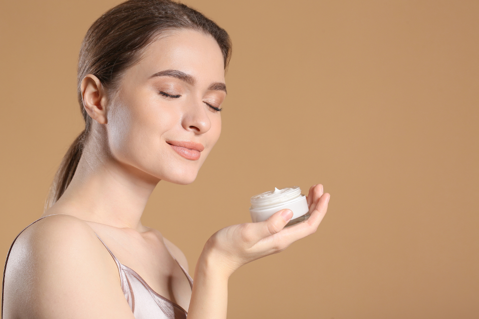 Woman with eyes closed smiles while holding an open white cosmetic jar against solid beige. — illustrating “Vichy Idealia Eyes and Serum Review” on Florence and Mary