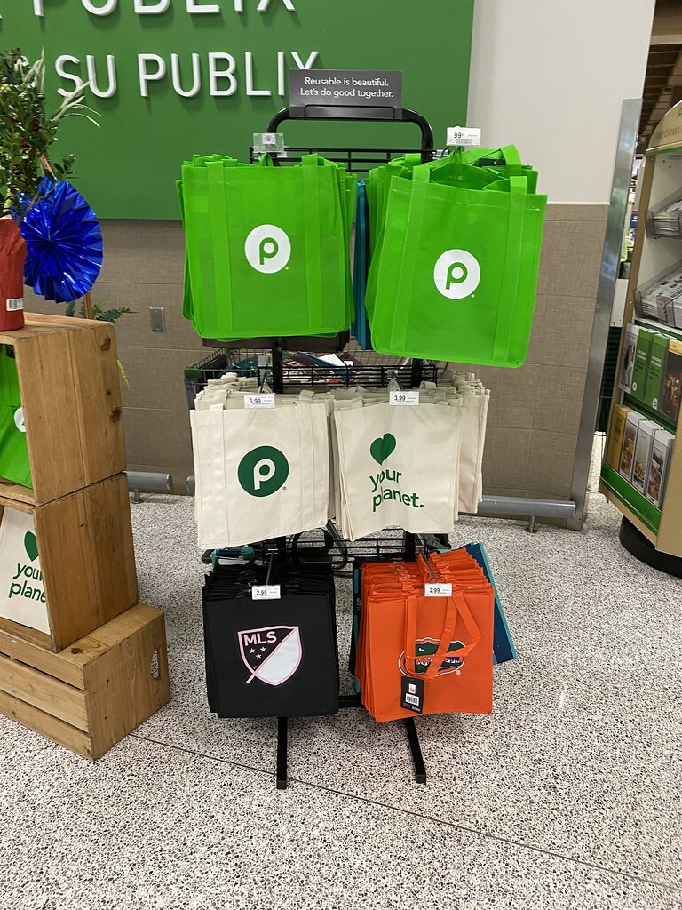Black metal grocery display rack holds bright green, white, black, and orange reusable shopping bags. — illustrating “Victoria Secrets US Haul” on Florence and Mary