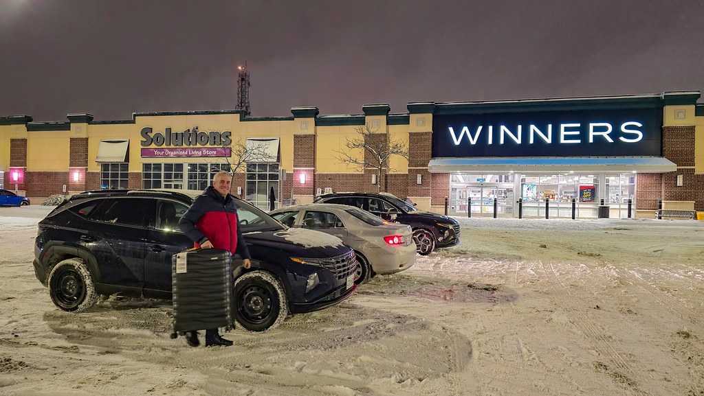 A man stands with a large black suitcase in a snowy parking lot at night before lit stores. — illustrating “Week 37 // What I'm Reading: The Longest Holiday” on Florence and Mary