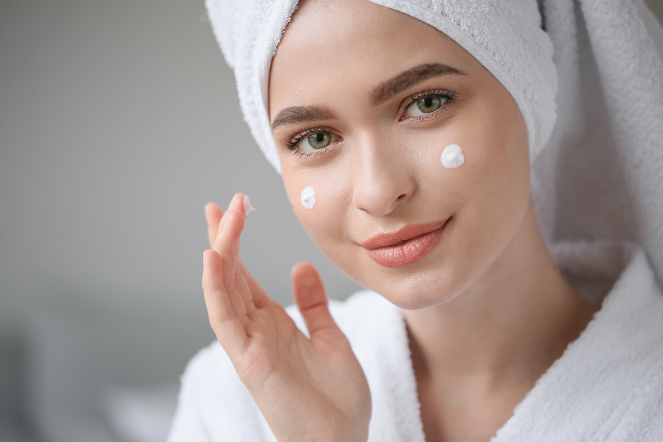 A smiling woman in a white bathrobe and towel applies white cream dots to her cheeks. — illustrating “What SPF are you using?” on Florence and Mary