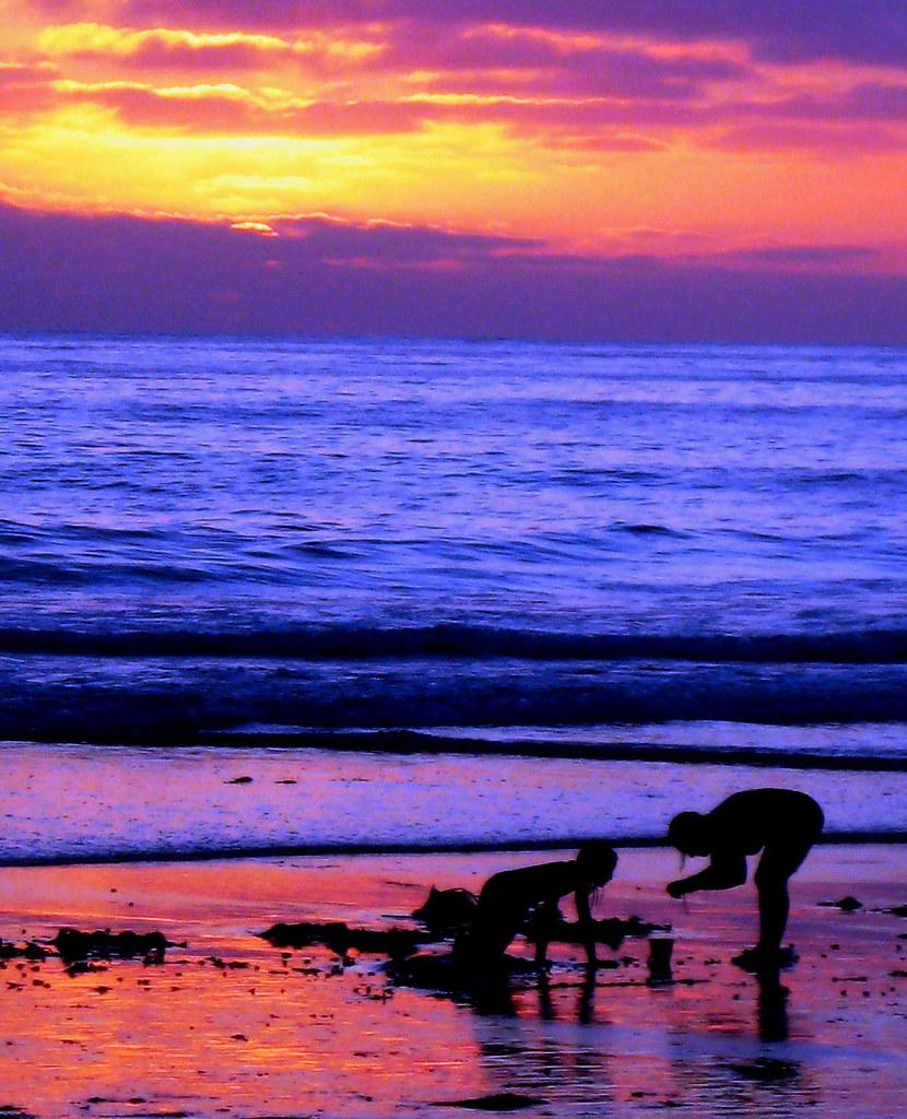 Two silhouetted children play on wet sand under a vivid orange-purple sunset by blue ocean. — illustrating “Whats in my beach bag” on Florence and Mary