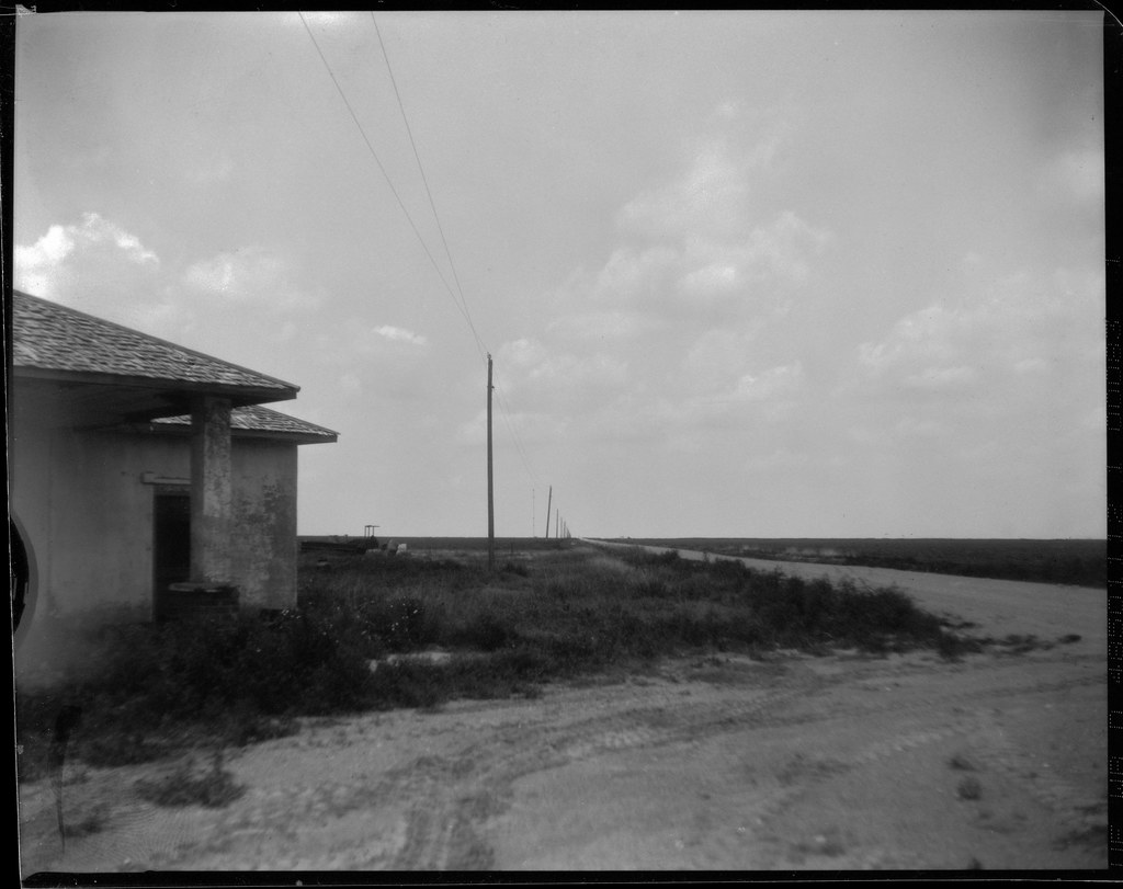 A black-and-white photo shows a small building and utility poles along a flat road under clouds. — illustrating “What's in my travel make up bag” on Florence and Mary
