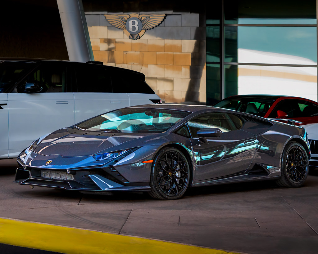 A dark grey metallic sports car parks before a building with a large winged Bentley emblem. — illustrating “What's Inside my hand luggage” on Florence and Mary