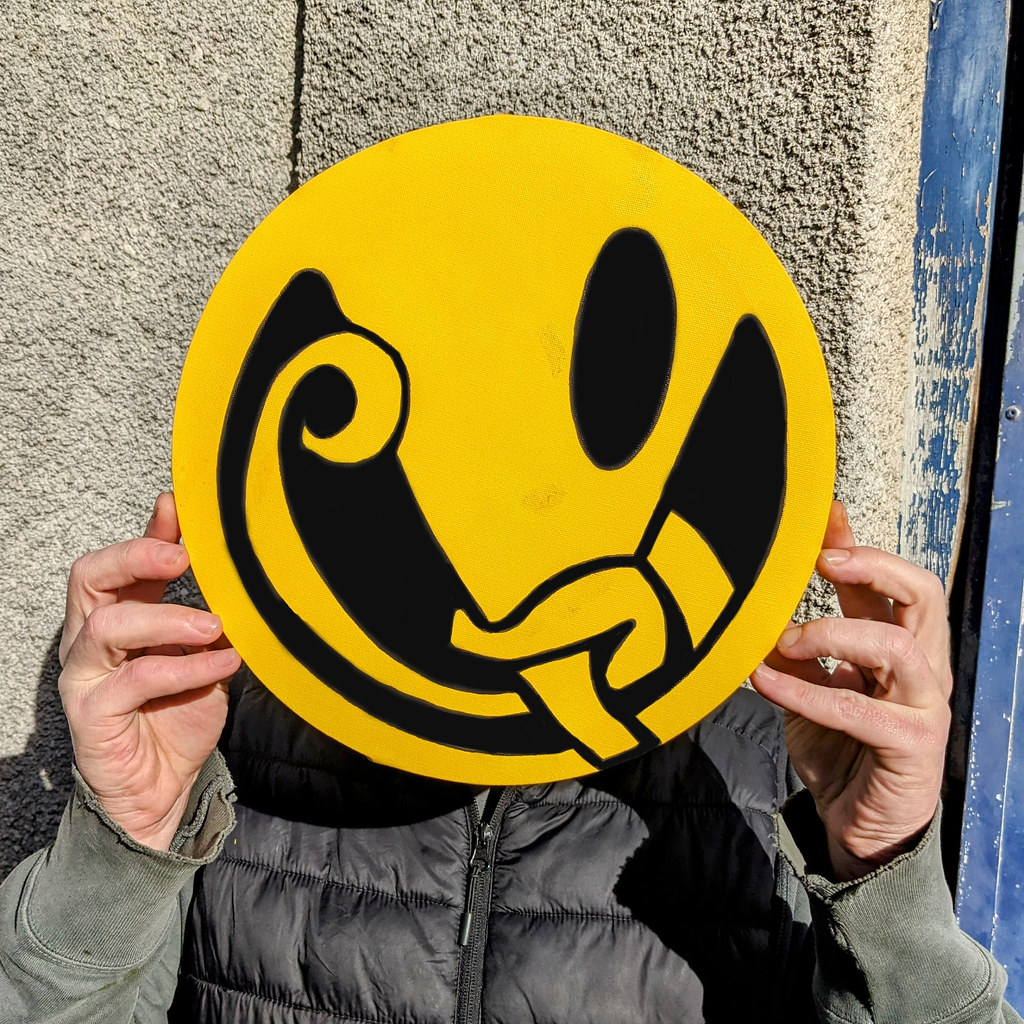 A person hides their face behind a large yellow circular canvas with a black abstract design. — illustrating “What's on my face // Cheering myself up” on Florence and Mary