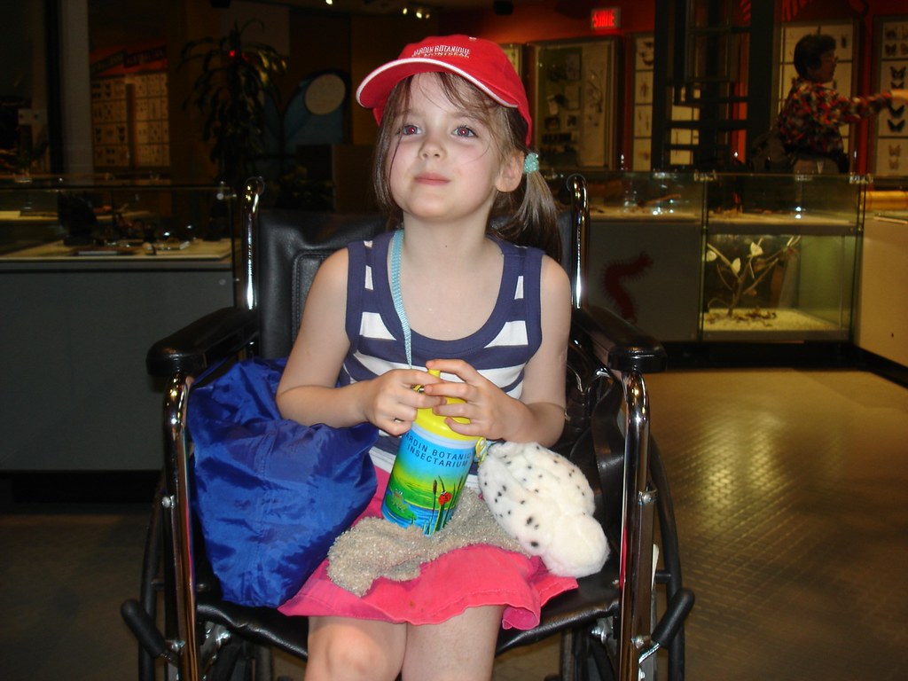 A young girl in a red cap and striped top sits in a wheelchair holding a plush toy. — illustrating “Birthday Days Out” on Florence and Mary