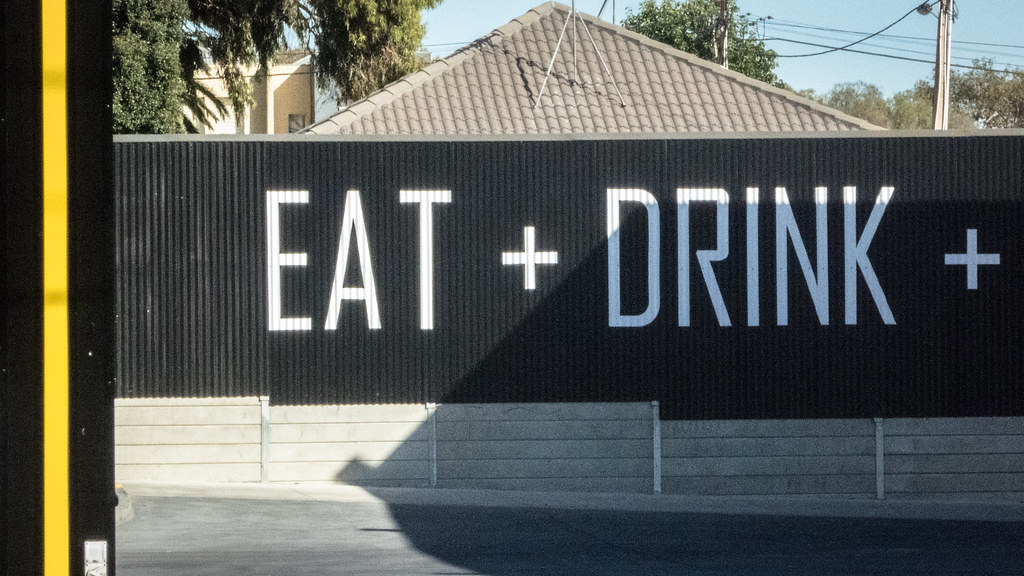 Large white letters spelling EAT + DRINK + appear on a black corrugated fence with trees behind. — illustrating “Yellow roses for the weekend” on Florence and Mary